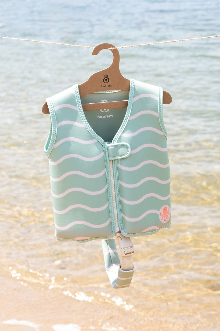 Babiem swim vest in mint green with white wave pattern and a small orange octopus motif, hanging on a branded wooden hanger against a calm seaside backdrop. The vest includes a white buckle strap visible at the bottom.