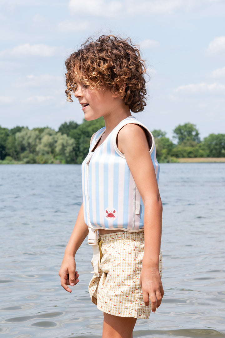 Young child wearing the Babiem Louis swim vest with blue and white stripes and a small red crab detail, standing at the edge of the water on a sunny day. The child looks to the side, enjoying a playful moment by the lake with trees in the background.