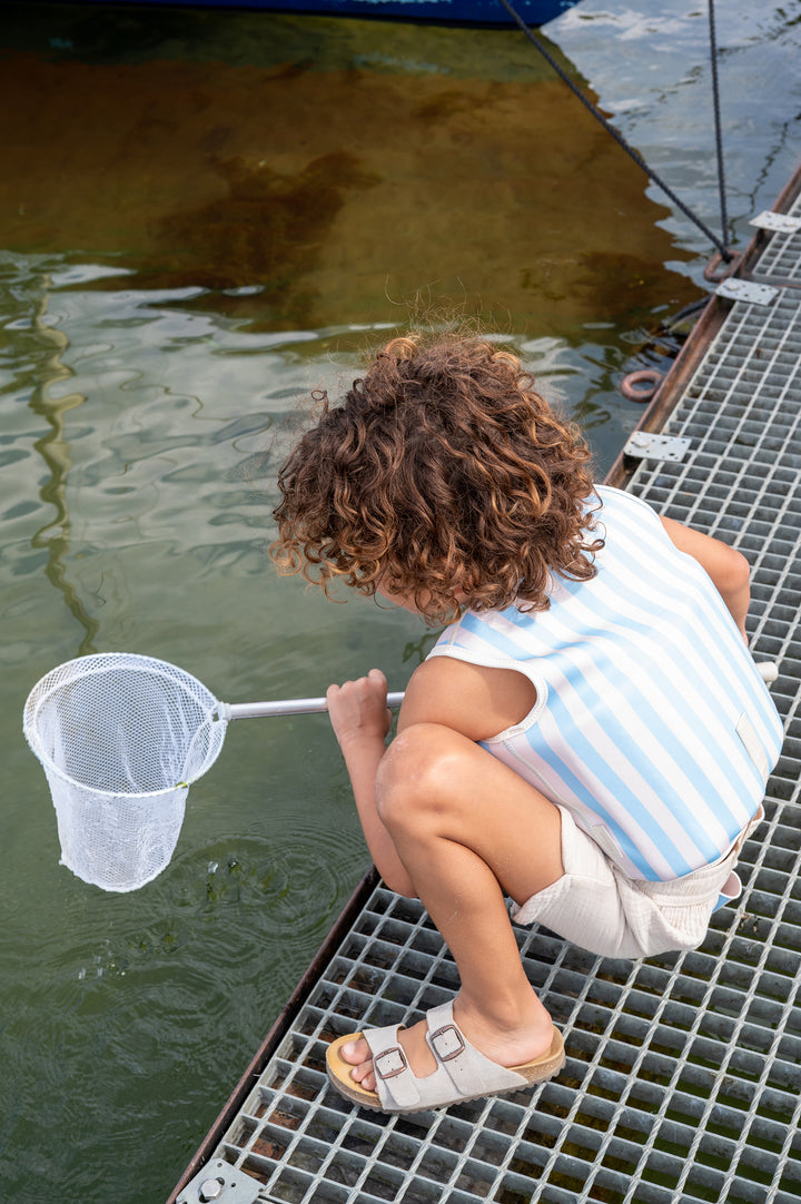 Child wearing the Babiem Louis swim vest with blue and white stripes, crouching on a pier while holding a small fishing net over the water. The swim vest features a red crab motif on the front and is secured with a white safety strap. The background shows calm water and part of a moored boat.