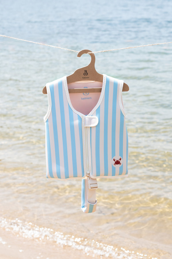 Babiem Louis swim vest in blue and white stripes with a red crab motif, hanging on a branded wooden hanger against a seaside backdrop. The vest is secured with a white buckle strap, and soft waves are visible in the background.