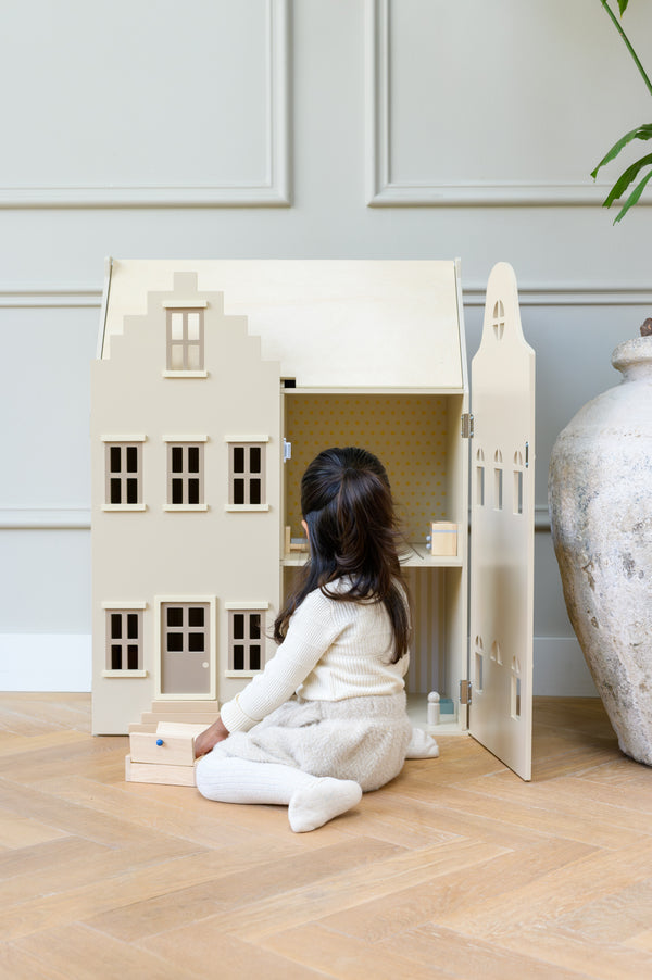 Young child in soft neutral clothing playing with the Babiem wooden dollhouse Lynn, a beige Dutch-style toy house with detailed windows and doors, in a bright and cozy interior.
