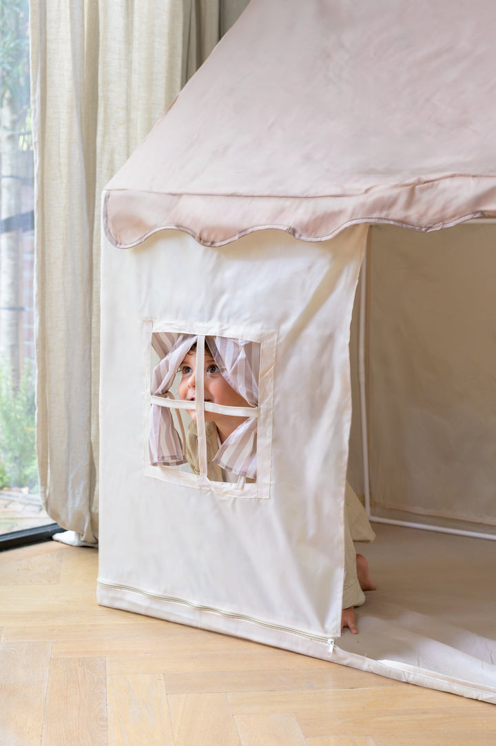 Smiling child peeking through the striped window curtains of the Babiem play tent Omar, a beige and taupe-colored fabric tent set up in a softly lit room with wooden flooring.