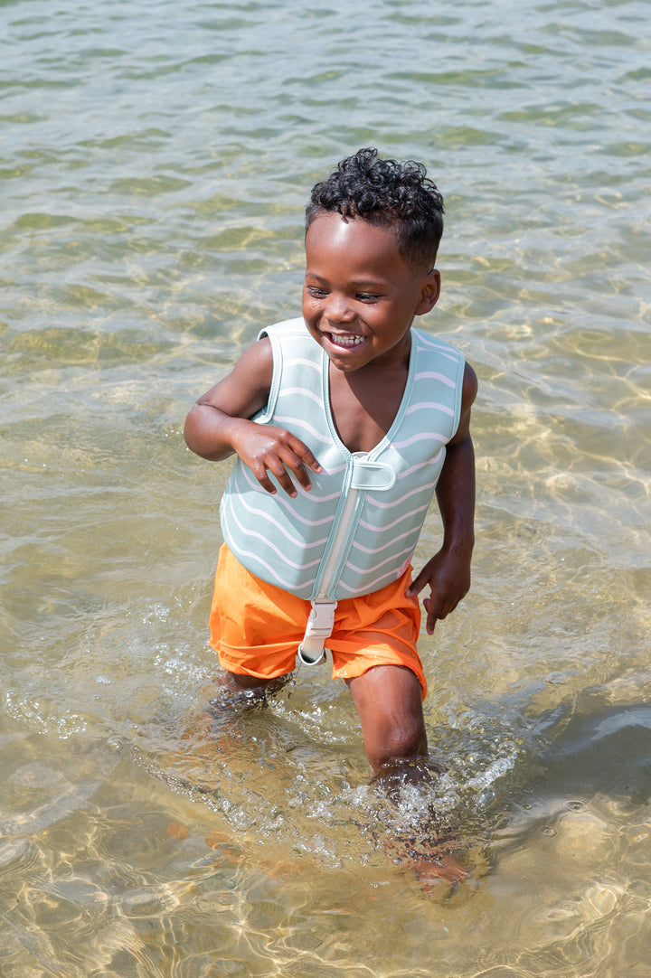 A smiling toddler in an aqua green swim vest with off-white wave pattern, running through shallow water on a sunny day.