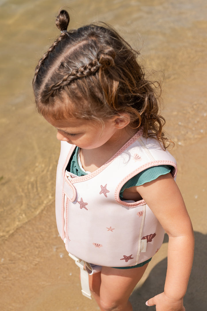 A young child with braided hair stands on the shoreline wearing swim vest Lily, decorated with starfish and shell illustrations in soft pink tones. The child looks down at the sand, with the clear water gently lapping nearby.