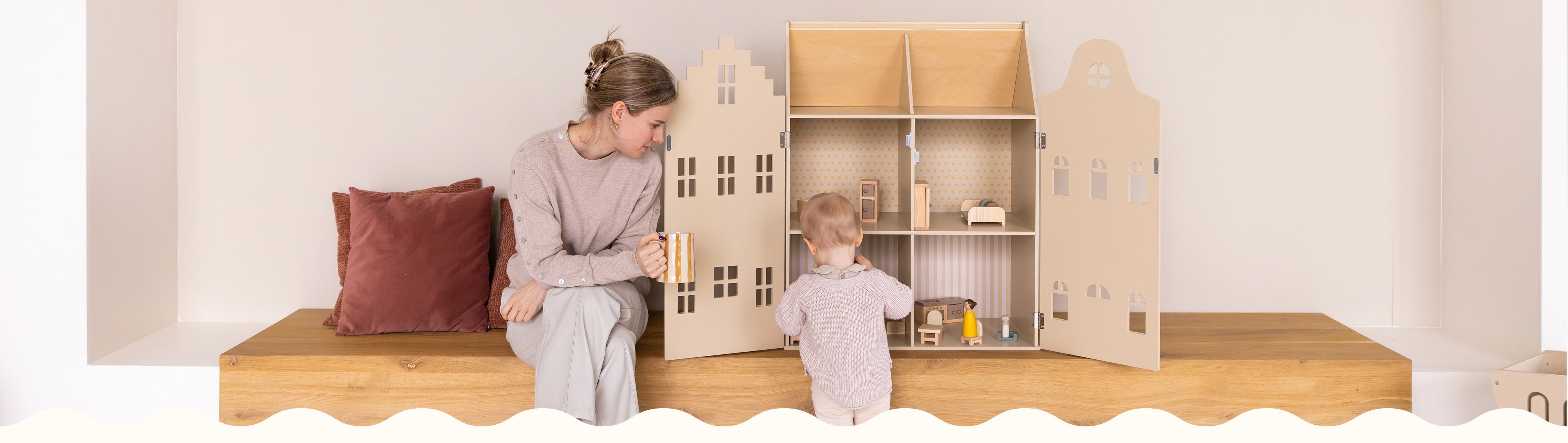 A cozy moment between a mother and daughter in a minimalistic playroom. The parent sits barefoot on a wooden bench with rust-colored pillows, holding a striped mug and watching the child play. The toddler stands at an open beige wooden dollhouse filled with miniature furniture and figures. The setting is calm and warm, with soft tones and natural textures throughout.