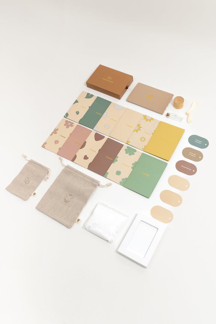 Top view of a baby milestone memory kit featuring a 1x storage box, 1x photo box (incl. 12x photo frame for each month), 1x clay and clay frame (14.7 x 10.4 x 2.5 cm), 2x canvas storage bag, 1x hair lock tube, 1x notebook and 6x hangtags (3 for your own interpretation). The items are designed in soft, earthy tones with a minimalistic and natural aesthetic.
