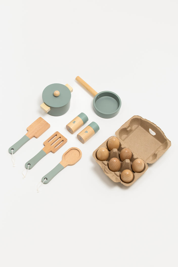 Top view of a wooden toy kitchen set including a pot with lid, frying pan, cooking utensils, salt and pepper shakers, and a cardboard egg carton with six wooden eggs. The items feature natural wood tones combined with muted green accents, designed for pretend play.