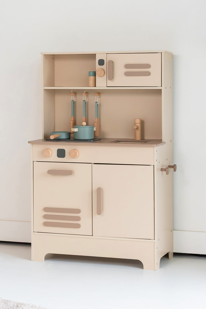 A minimalist wooden toy play kitchen in beige tones stands against a white wall. On the countertop are two blue-green pans with wooden handles and three wooden spatulas hanging on hooks. To the right is a wooden tap above a built-in sink. A cupboard and a pot are visible at the top. The design is clean and modern, with rounded corners and calm colours that fit into a Scandinavian interior.