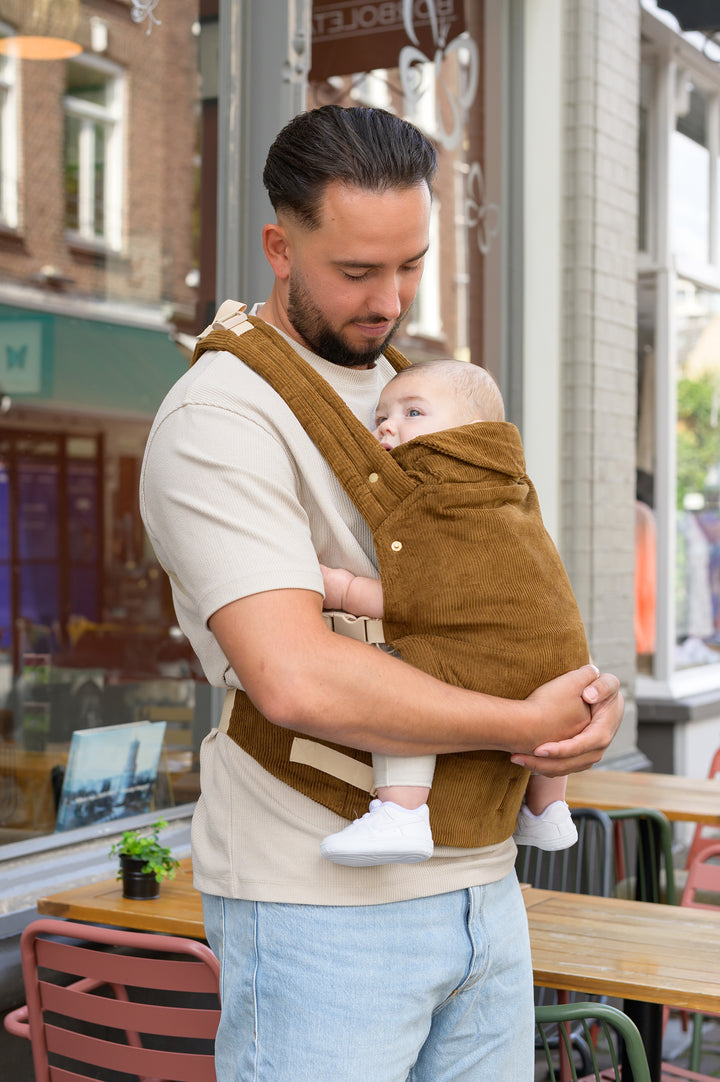 A father standing outside in the city, holding his baby close in our brown corduroy Babiem baby carrier Luca. He looks down warmly at the baby, creating a cozy and intimate moment together.