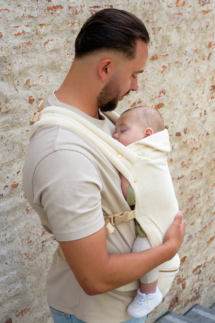 A father gently holding his sleeping baby close in a soft Babiem baby carrier. They are standing near a rustic brick wall, sharing a quiet and tender moment together.