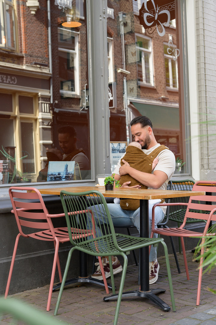 A father sitting at an outdoor café table, holding his baby close in our brown corduroy Babiem baby carrier Luca. He looks down affectionately at the baby, enjoying a quiet and cozy moment together.