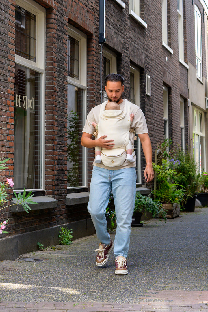 A father walking down a quiet street holding his baby in a soft Babiem baby carrier. He is wearing casual jeans and sneakers, keeping his baby close and comfortable during the stroll.