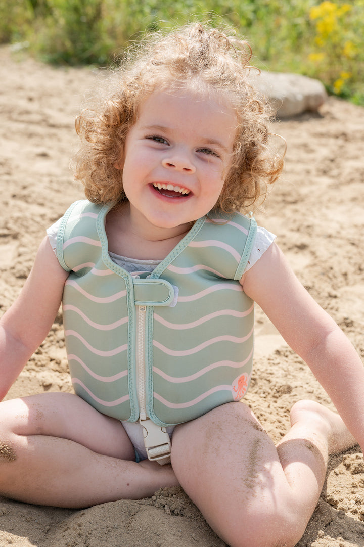 A smiling young girl sitting on the beach in a mint green swim vest with wavy off-white lines, named Charlie. She’s playing happily in the sand.