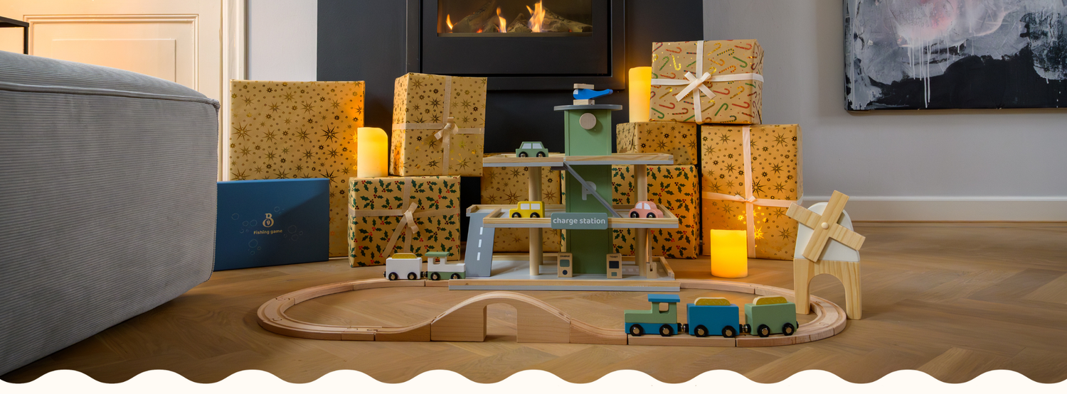 A warm holiday scene featuring Babiem wooden toys, including a train set, a windmill and a play garage, surrounded by wrapped gifts and soft candlelight in front of a fireplace.