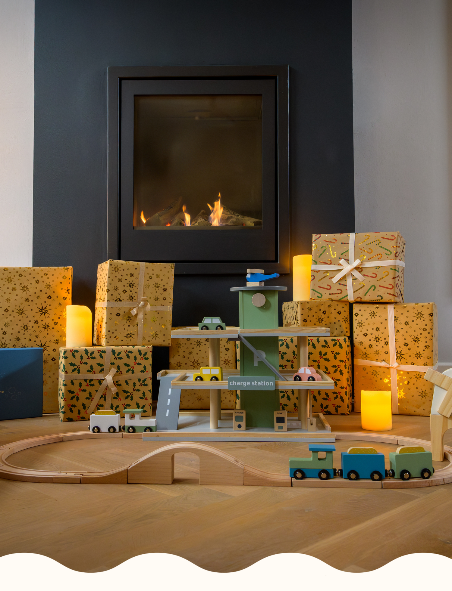 A warm holiday scene featuring Babiem wooden toys, including a train set, a windmill and a play garage, surrounded by wrapped gifts and soft candlelight in front of a fireplace.
