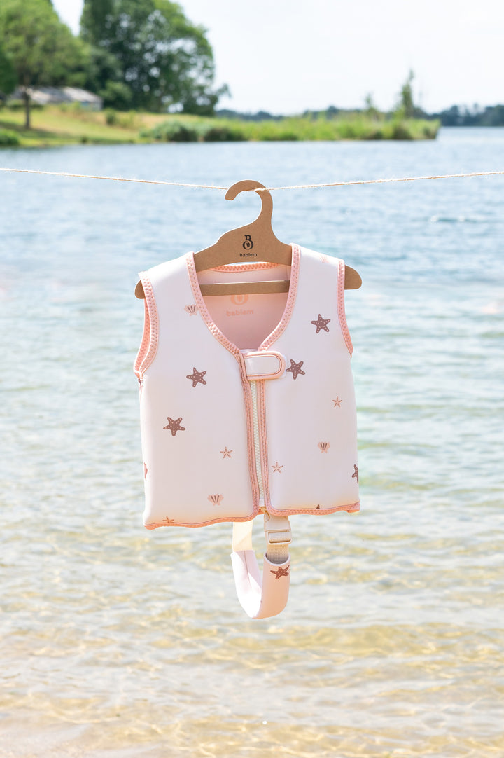 A close-up of the Lily swim vest from Babiem, decorated with starfish and seashells in soft pink tones. The vest hangs neatly on a branded wooden hanger over clear lake water with a green shoreline and blue sky in the background.