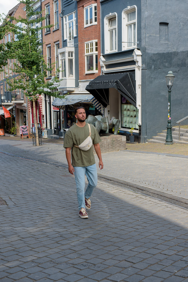 A man walking through a city street wearing a Babiem fanny pack, which is part of the Babiem baby carrier Chloé. He is dressed casually in jeans and a green T-shirt, using the fanny pack for practical everyday storage.