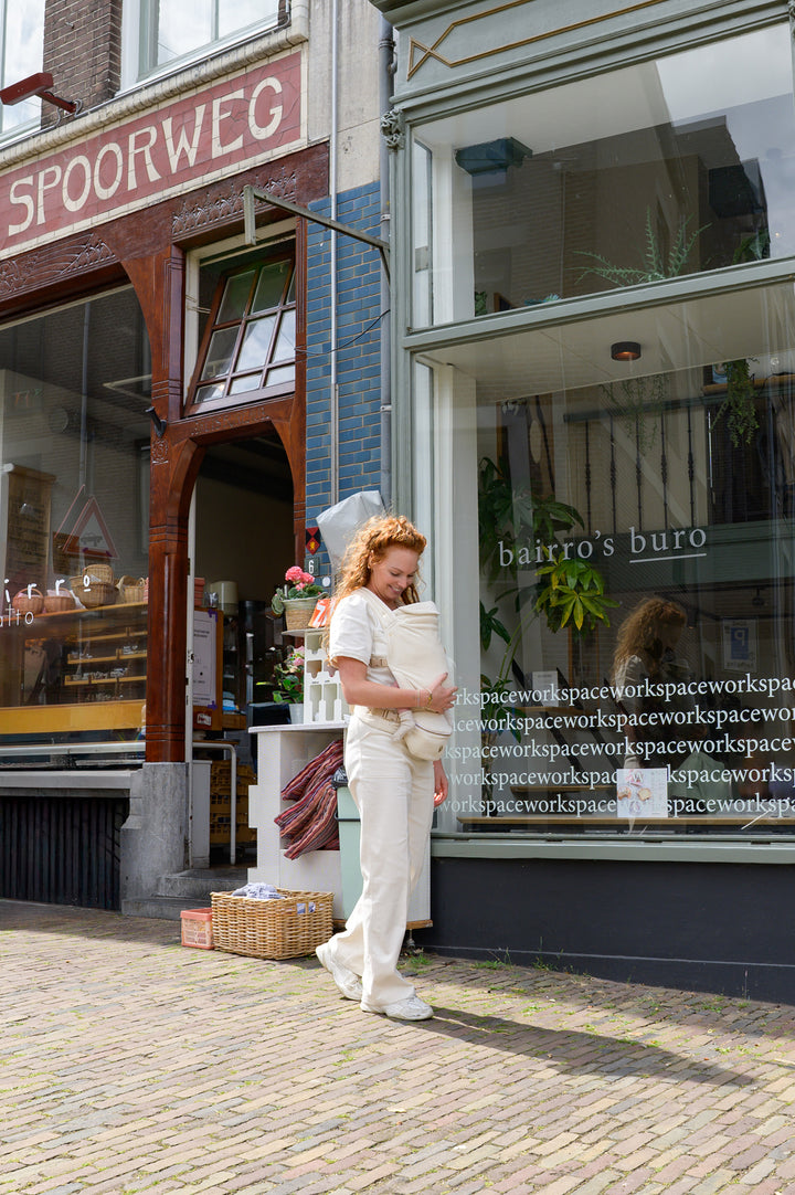 A smiling mother stands in front of a shop, holding her baby close in a soft Babiem baby carrier. She looks down lovingly at her baby while enjoying a peaceful moment outdoors.