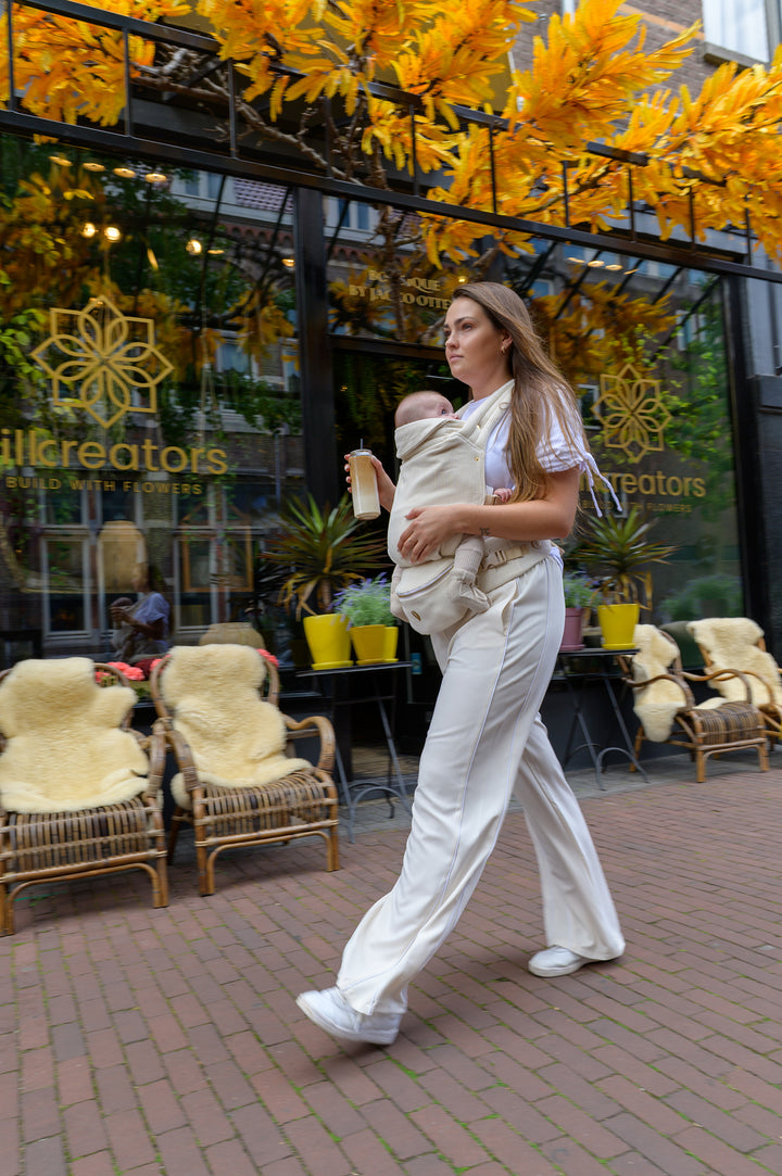 A mother walking past a flower shop holding her baby close in a soft Babiem baby carrier. She carries a coffee to go in her hand, enjoying a calm moment together during their stroll.