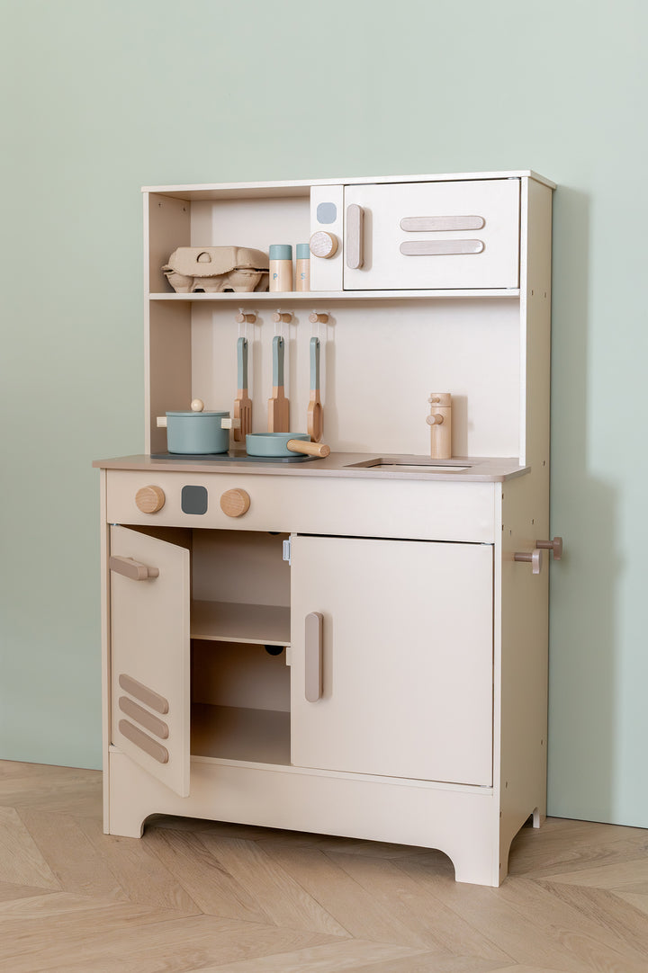 Play kitchen Noor by Babiem with open cabinet doors revealing storage space. On the countertop are pastel-colored pots, utensils, and a wooden faucet; on the shelf are a carton of eggs and spice containers.