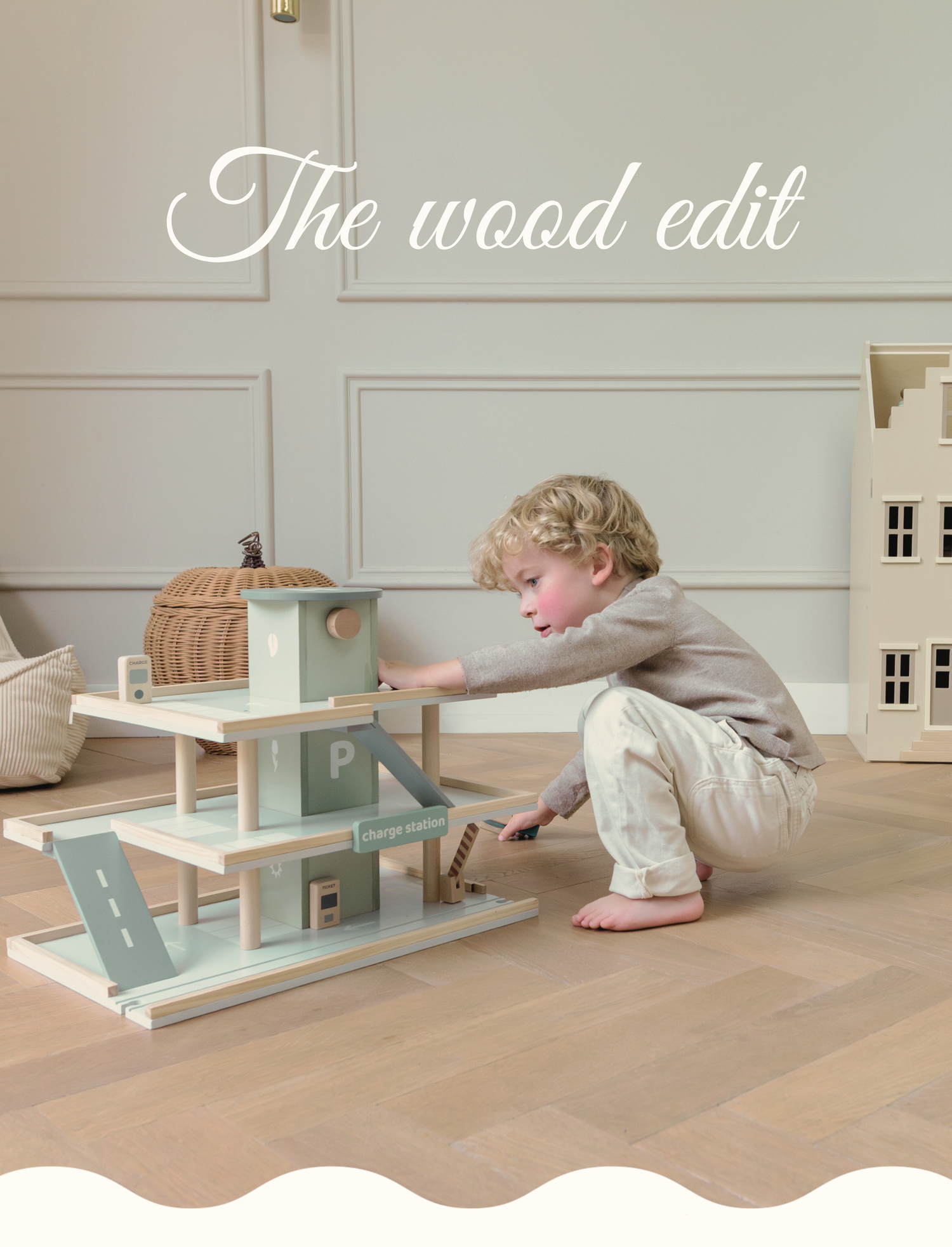 A child playing with a minimalist wooden toy garage on a light wood floor, part of the wood edit collection for Babiem. In the background, there is a wooden dollhouse and a wicker basket. The image features a soft, natural aesthetic with the text "The wood edit" in white script.