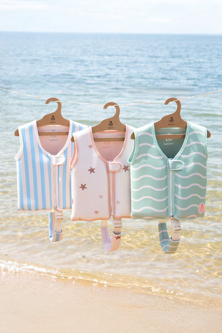 Three children's swim vests in pastel tones light blue with stripes and a crab, pink with sea creatures, and mint with wave patterns and an octopus, hanging neatly on wooden hangers above the shoreline, with the ocean in the background.
