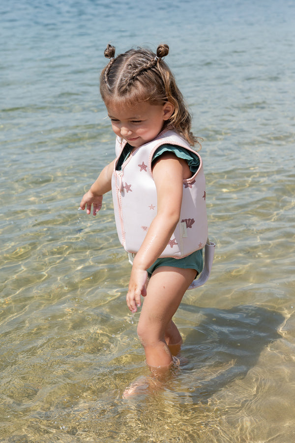 A young child with braided hair stands in clear, shallow water while wearing the Babiem swim vest Lily in soft pink with sea star and shell illustrations. The child is looking down with a gentle smile, wearing a green swimsuit underneath.
