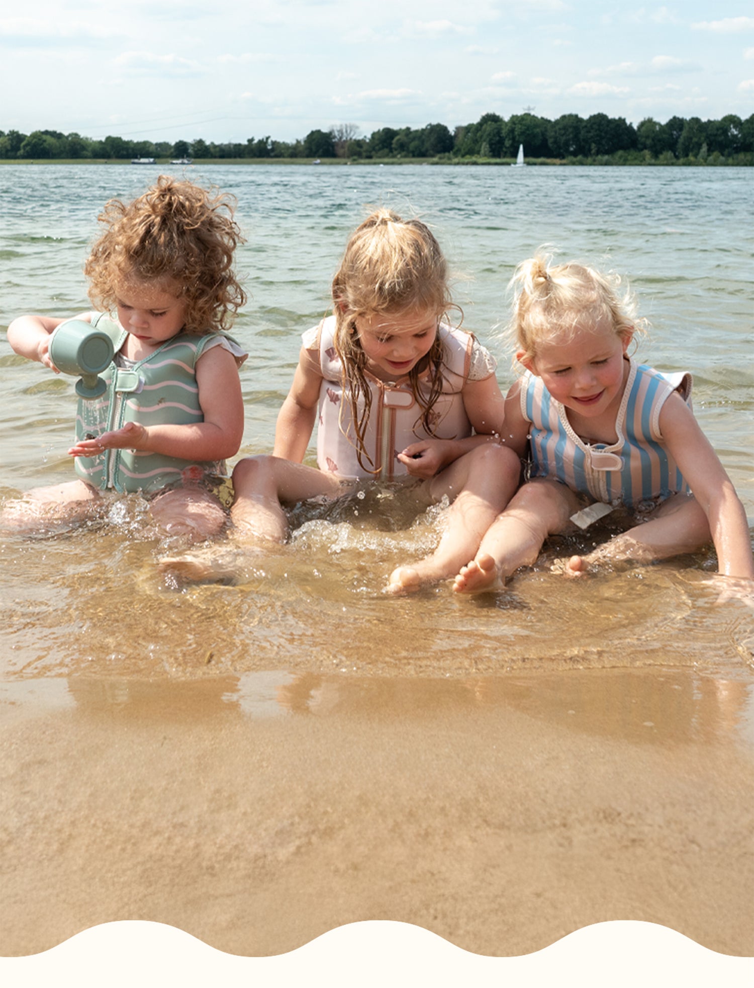 Three toddlers sit at the shoreline, playing with water and sand while wearing Babiem swim vests in pastel green, pink, and blue. One child pours water from a small watering can, while the others watch and splash in the shallow water.