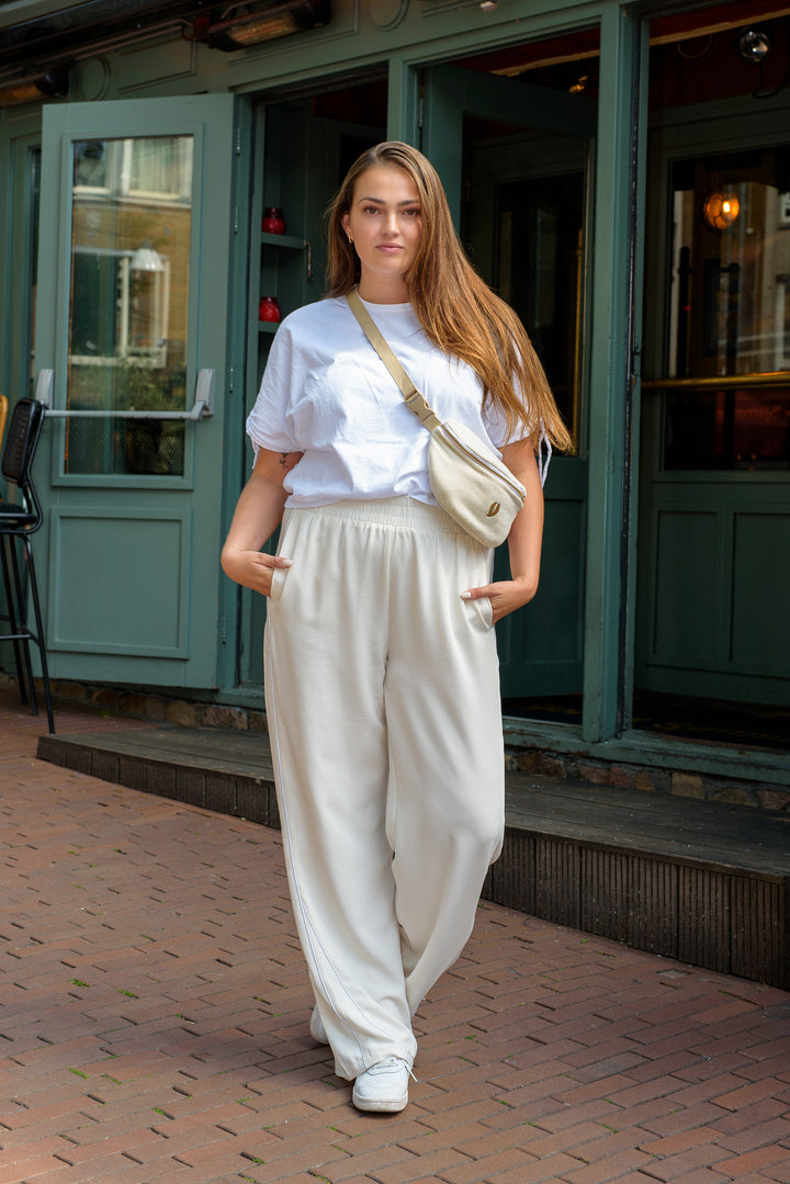 A woman standing outside in the city wearing a off-white velvet Babiem fanny pack across her chest. The fanny pack, part of the Babiem baby carrier Olivia, is styled casually with her white outfit for everyday use.