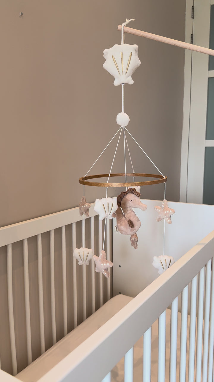 A soothing Babiem music mobile Zoë hanging above a baby’s crib, featuring soft fabric sea-themed shapes like a seahorse, shells, and stars in gentle beige and white tones. The mobile plays calming music, creating a peaceful and dreamy nursery atmosphere.