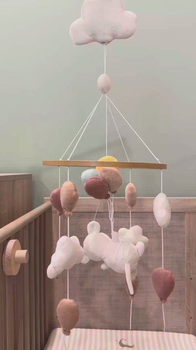 Musical baby mobile Sam from Babiem with plush white clouds and multi-colored balloons in earth tones, hanging from a wooden hoop above a light wood crib and playing music.