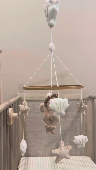 Babiem baby mobile featuring plush ocean-themed toys including a starfish, and seashells in soft neutral colors,  above a light wood crib, playing music.
